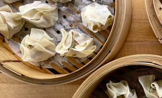 Three bamboo steamers filled with uncooked dumplings, arranged on a wooden surface. The dumplings are wrapped in thin pastry and placed on greaseproof paper inside the steamers.