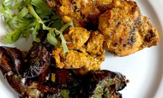 Grilled pieces of seasoned chicken served with roasted aubergine garnished with herbs and a small side of fresh rocket on a white plate.