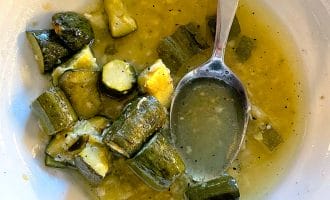 A white bowl containing chunks of cooked courgette in a yellow-green broth, with a metal spoon resting in the bowl. Some broth and vegetable pieces are scattered around.