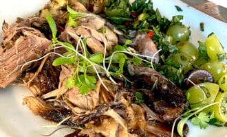 A plate of shredded cooked meat with microgreens, roasted vegetables, sliced green grapes, and fresh herbs, served on a white dish on a wooden table. A knife rests on the plate’s edge.