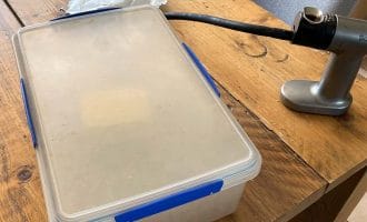 A plastic container with a blue catch sits on a wooden table beside a silver handheld kitchen tool with a black hose attached. A piece of crumpled paper is also on the table near the container.