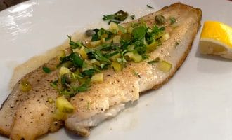 A cooked fish fillet garnished with chopped herbs and capers is served on a white plate with a lemon wedge on the side. A hand holds the plate, and part of a patterned garment is visible in the background.