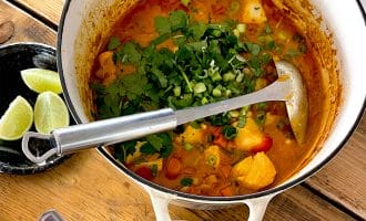 A pot of fish stew with chunks of fish, vegetables, and fresh herbs sits on a wooden table next to a bowl of white rice and a plate with lime wedges. A ladle rests inside the pot.