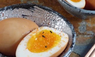 A ceramic bowl with a halved soft-boiled egg, seasoned with spices and black sesame seeds. Another bowl contains whole and halved eggs. Chopsticks are placed nearby on a stone surface.