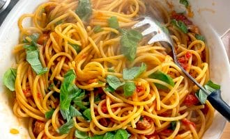 A bowl of spaghetti with tomato sauce, fresh basil leaves, and a pasta serving spoon, sitting on a wooden surface. Steam rises from the pasta, indicating it is freshly cooked.