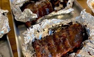 Barbecued ribs covered in sauce, partially wrapped in aluminium foil, served on metal trays on a wooden table. The ribs appear juicy and well-cooked, ready to be eaten.
