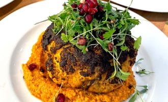 A whole roasted cauliflower sits on a bed of orange sauce, garnished with fresh microgreens and pomegranate seeds, served on a white plate.
