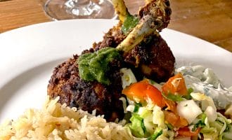 A plate of spiced chicken drumsticks topped with green sauce, served with white rice, chopped vegetable salad, and a side of creamy sauce, with a wine glass in the background.
