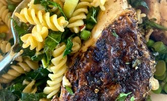 Close-up of a plate with seasoned grilled chicken breast, rotini pasta, leafy greens, peas, and vegetables, all mixed together. A fork is partially visible on the left side of the dish.