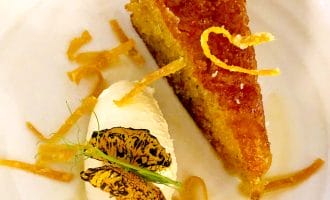 A slice of golden syrup cake garnished with thin orange zest strips, served with a scoop of vanilla ice cream and caramelised orange slices on a white plate.