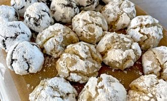A container lined with brown baking parchment holds rows of crinkle biscuits. The biscuits are covered in icing sugar; some are chocolate and others are light-coloured, suggesting different flavours.