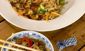 A large white plate and a small patterned bowl filled with stir-fried noodles, vegetables, and spring onions sit on a wooden table. Chopsticks rest on the bowl, with a chopstick holder nearby.