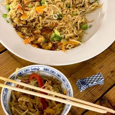 A large white plate and a small patterned bowl filled with stir-fried noodles, vegetables, and spring onions sit on a wooden table. Chopsticks rest on the bowl, with a chopstick holder nearby.