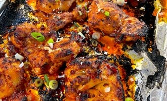 Charred spicy baked chicken wings on a foil-lined tray, garnished with sliced spring onions and sesame seeds. The wings are coated in a red sauce, with crispy edges and visible bits of seasoning.