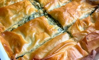 A close-up of golden-brown, flaky spanakopita cut into rectangles in a white baking dish, with crispy filo pastry visible on top and edges.