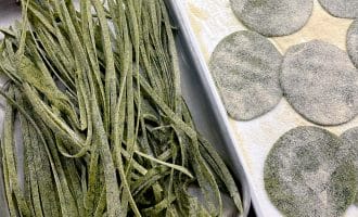 Fresh green pasta dough cut into fettuccine strips sits in one tray, while round green ravioli wrappers dusted with flour are arranged on another tray. Both trays are on a wooden surface.