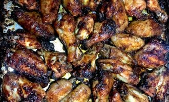 A close-up view of a baking tray filled with numerous baked chicken wings, golden-brown and caramelised, with crispy, charred edges.