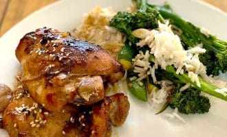 A plate of glazed chicken thighs sprinkled with sesame seeds, served with white rice and sautéed green vegetables, including tenderstem broccoli, on a white plate on a wooden table.
