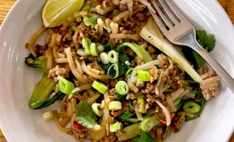 A white bowl filled with stir-fried noodles, minced meat, sliced spring onions, coriander, cucumber, and a lime wedge, with a fork resting on the edge of the bowl, on a wooden table.