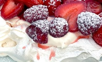 A close-up of a pavlova dessert topped with whipped cream, fresh strawberries, raspberries, and a dusting of icing sugar on a white plate.