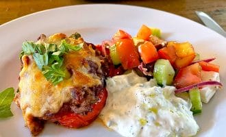 A white plate with a serving of baked dish topped with melted cheese and herbs, a scoop of creamy tzatziki, and a colourful Greek salad with tomatoes, cucumber, red onion, and feta cheese. A baking dish is visible in the background.