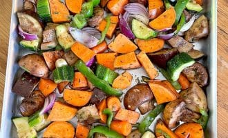 A baking tray filled with chopped mixed vegetables including sweet potatoes, courgette, aubergine, carrots, green pepper, red onion, mushrooms, and whole garlic cloves, ready for roasting.