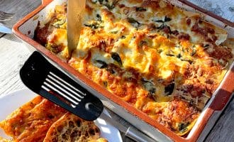 A baked lasagne in a rectangular dish with a knife inserted, surrounded by golden melted cheese and herbs. A plate with slices of crusty bread and a black spatula sits nearby on a sunlit wooden table.