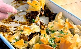 A person serves a portion of a baked dish with golden, crispy filo pastry on top, revealing a rich, dark filling underneath in a white rectangular baking dish. Parsley is sprinkled on the pastry.