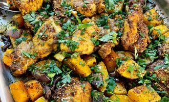 A tray of roasted chicken pieces and diced vegetables, including yellow courgette, onions, and herbs, garnished with chopped fresh parsley and spices. The dish appears golden-brown and flavourful.