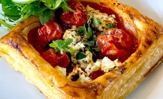 A golden puff pastry square filled with roasted cherry tomatoes, crumbled cheese, and herbs, served on a white plate with a side of fresh rocket.