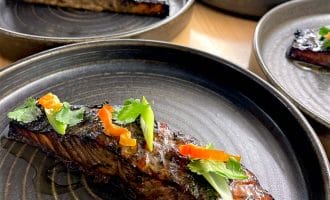 A close-up of a glazed salmon fillet garnished with sliced red peppers, spring onions, and coriander, served on a dark ceramic plate with more plated salmon portions in the background.
