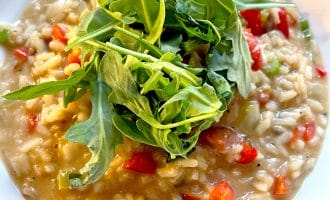 A close-up of creamy risotto with diced red peppers, garnished with a handful of fresh rocket, served in a white bowl.
