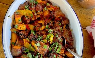 A white oval dish filled with a colourful roasted vegetable medley, including sweet potatoes, peppers, and mushrooms, garnished with chopped herbs. A spoon rests in the dish, and salt and chilli flakes are nearby.