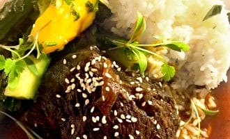 Braised beef topped with sesame seeds, served with white rice, sautéed green vegetables, and slices of yellow pepper, garnished with fresh herbs on a white plate.