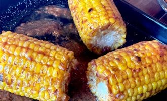 Three pieces of grilled corn on the cob, coated with seasoning and butter, sit on a black tray with melted butter and spices pooling around them.