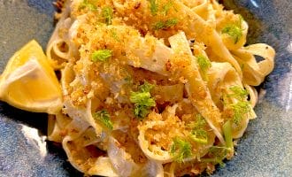 A bowl of fettuccine pasta topped with golden breadcrumbs and fresh herbs, garnished with fennel fronds and served with a lemon wedge on the side.