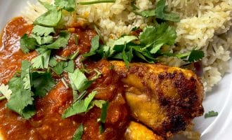 A plate of chicken curry with rich red sauce, served alongside seasoned rice and garnished with fresh coriander leaves.