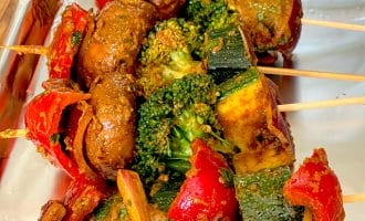 Vegetable skewers with mushrooms, broccoli, courgette, red peppers, and onions covered in seasoning, arranged on a foil-lined tray.