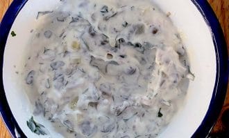 A white bowl with a blue rim filled with a creamy mixture containing chopped herbs and vegetables, sitting on a wooden surface.