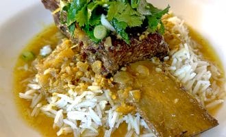 A braised beef short rib served over white rice, topped with fresh coriander and chopped spring onions, with a golden sauce poured around the dish on a white plate.