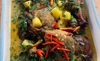 A roasting dish filled with roasted chicken pieces in a yellow curry sauce, garnished with pineapple chunks, chopped coriander, and sliced red chilli peppers.
