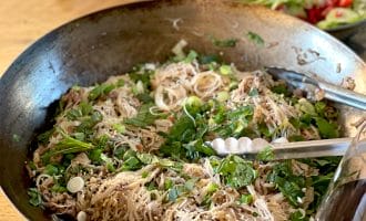 A large pan filled with cooked rice noodles, chopped spring onions, herbs, and sesame seeds, with metal tongs resting on the side. In the background, there is a bowl of mixed salad with tomatoes and cucumbers.