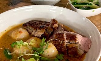 A bowl of red curry with sliced duck, round vegetables, and herbs, served with a side of white rice in a patterned bowl and a small dish of cucumber salad on a wooden table.