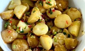 A white bowl filled with halved baby potatoes mixed with chopped spring onions, red chilli, and herbs, placed on a wooden surface.