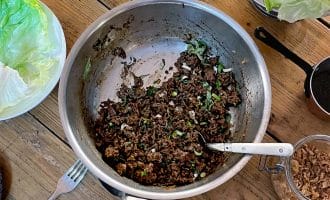 A metal pan with cooked minced meat and chopped spring onions sits on a wooden table, surrounded by lettuce leaves, a bowl of rice, cutlery, and small dishes with various toppings.