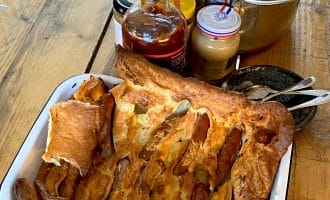 A tray of toad in the hole (sausages baked in Yorkshire pudding batter) sits on a wooden table next to a pot of mashed potato, jars of condiments, and some serving utensils.