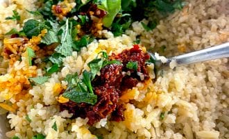 A close-up of cooked rice mixed with fresh chopped herbs and a spoonful of red sauce, stirred together in a white bowl with a metal spoon.