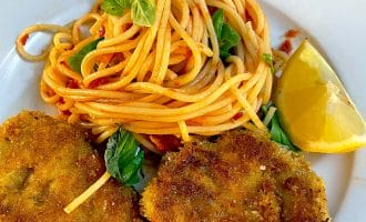 A plate with a serving of spaghetti in tomato sauce, garnished with basil leaves, two pieces of breaded fried meat, and a lemon wedge on the side.