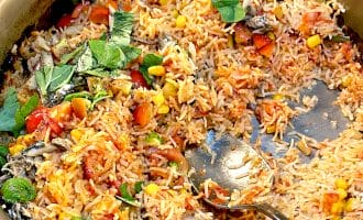 A large metal pan filled with mixed rice, sweetcorn, tomatoes, leafy herbs, and small pieces of fish. A serving spoon rests in the rice, which appears freshly cooked and partially served.