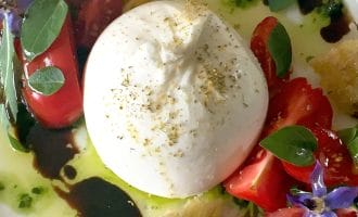 A plate with a ball of fresh burrata cheese, cherry tomato halves, toasted bread pieces, edible purple flowers, green leaves, olive oil, and a drizzle of balsamic glaze.